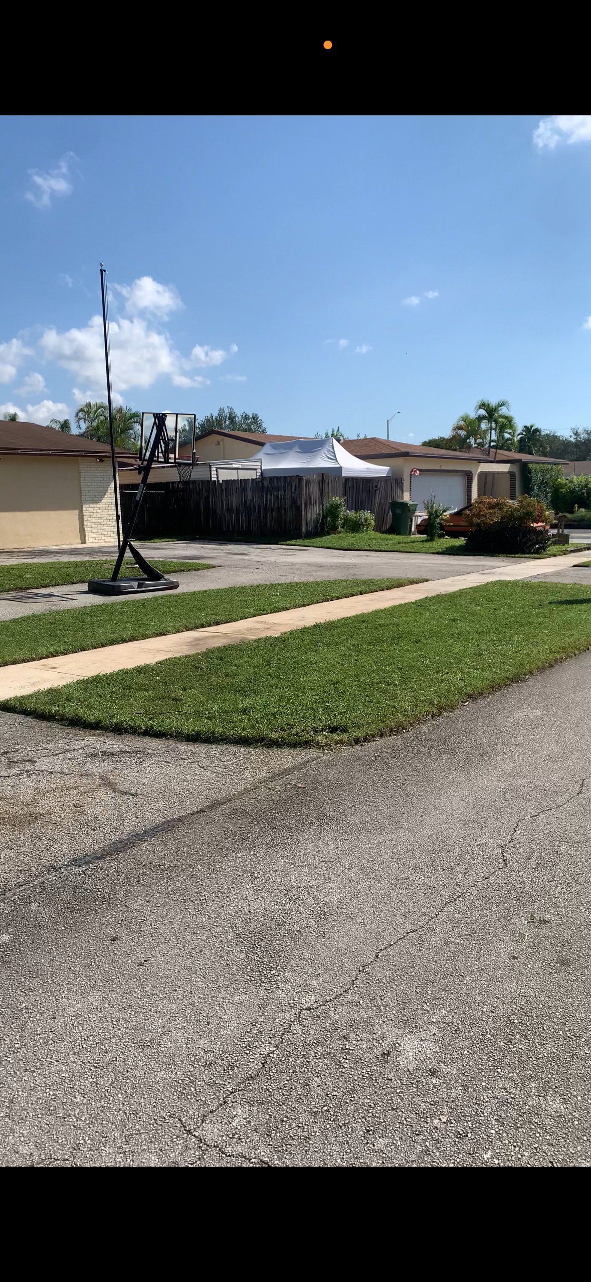 Suburban residential street with single-story homes, manicured lawn, palm trees, and construction crane in driveway under clear blue sky